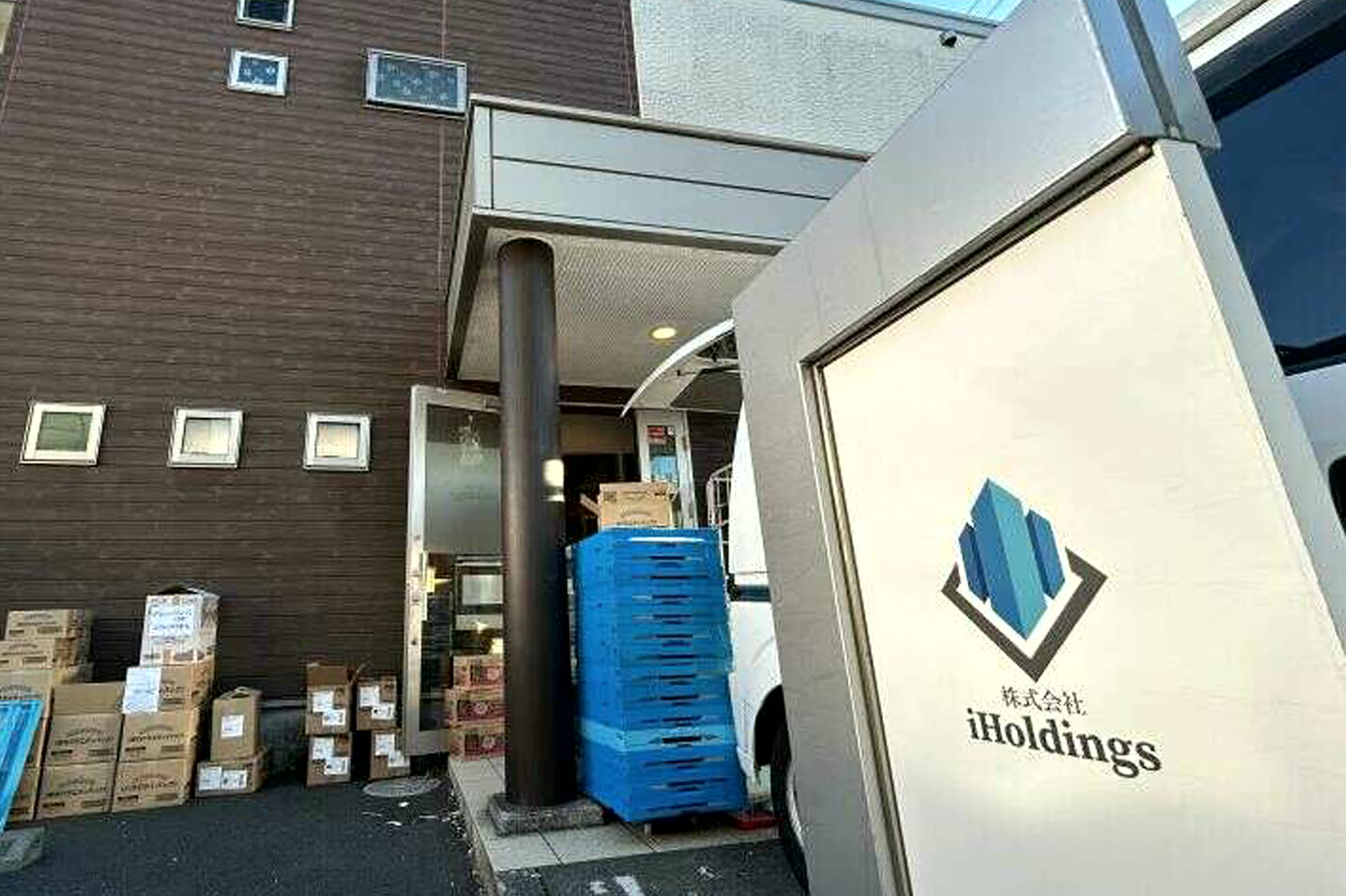 iHoldingsの社屋前に停まる、ロゴ入りの配送車と積み上げられた段ボール箱